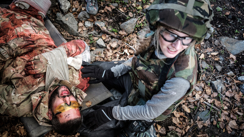 Military medic attending to wounded soldier on rocky ground during combat scenario.