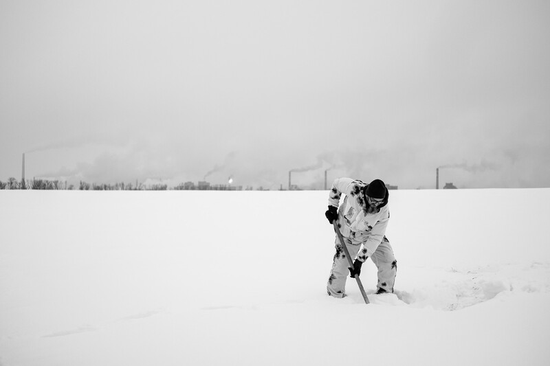 Person in winter camouflage crouching in deep snow under overcast sky.