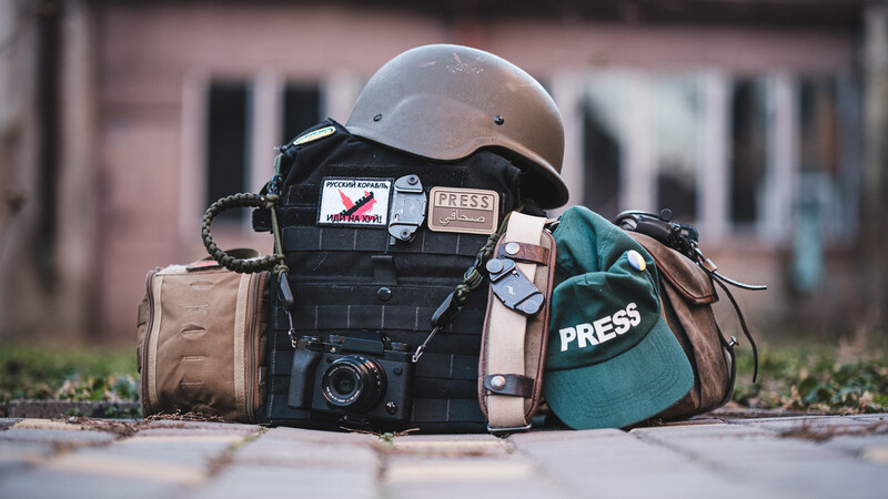 Professional photographer's gear arranged on pavement including camera body, white helmet, press vests, and tan carrying bag.