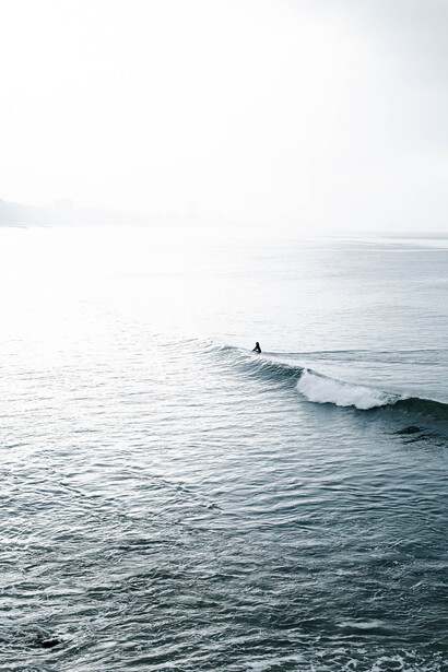 Solitary surfer paddling toward a rolling wave under overcast sky.