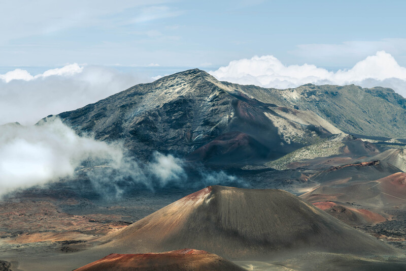 Volcanic cones with reddish-brown slopes in foreground, larger mountain shrouded in mist in background under blue sky.