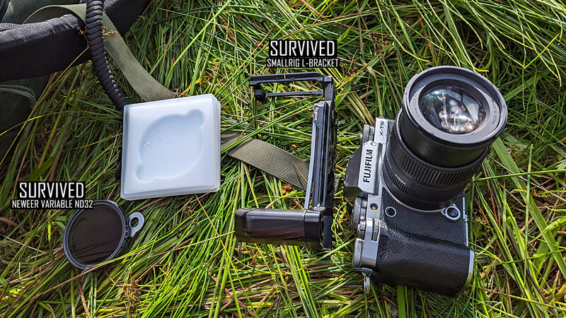 Photography gear laid out on grass including a camera with lens, diffuser, lens hood, and other accessories with survival labels.