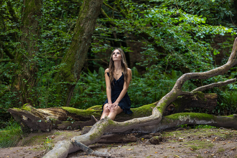 Woman in black dress kneeling on moss-covered fallen log in dense forest, gazing upward.