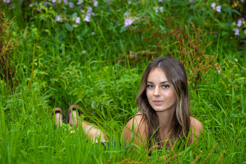 Young woman with long dark hair sitting in tall green grass, looking toward camera with neutral expression.