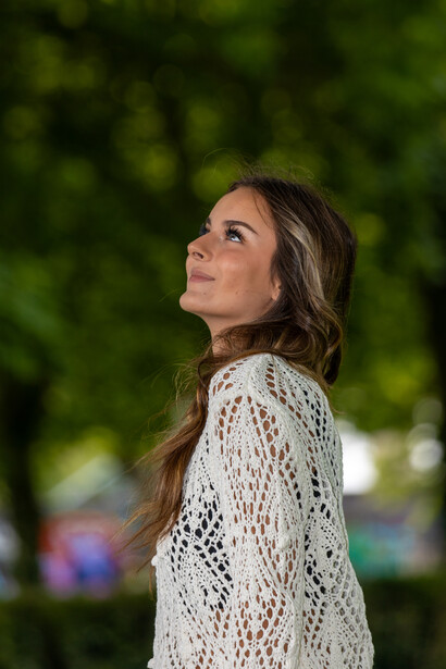 Woman in white crochet cardigan looking upward outdoors with blurred green foliage background.