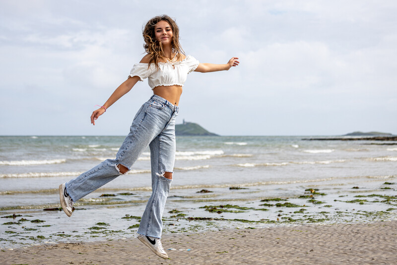 Woman jumping on a beach with arms outstretched, wearing a white crop top and distressed jeans.