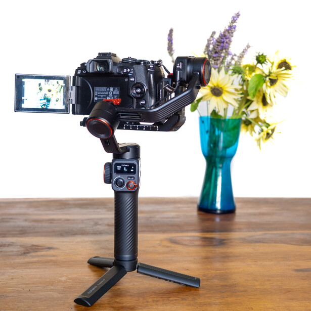 Cinema camera mounted on three-axis gimbal stabilizer with monitor displaying live feed and vase of flowers alongside.