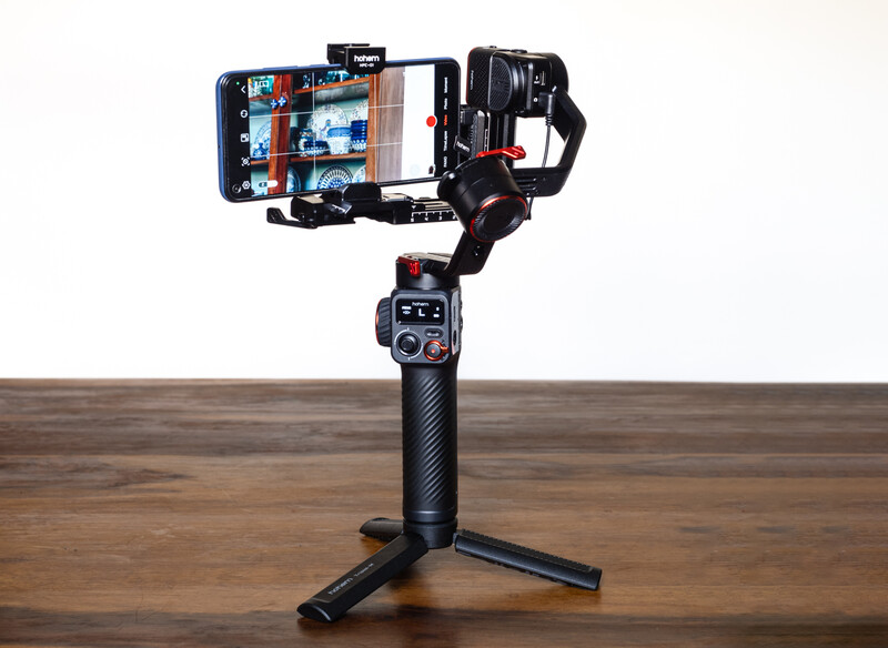 Three-axis smartphone gimbal mounted on a tripod stand with a phone attached to the top arm.