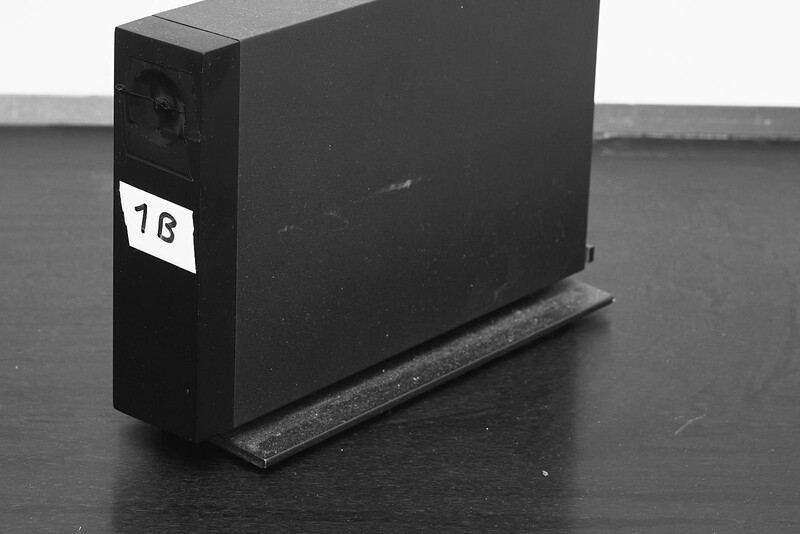 Black rectangular equipment box with white label marked "1B" sitting on a dark stand against a gray surface.