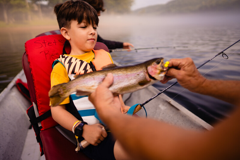 Young boy in red life jacket holding a freshly caught fish while fishing from a boat.