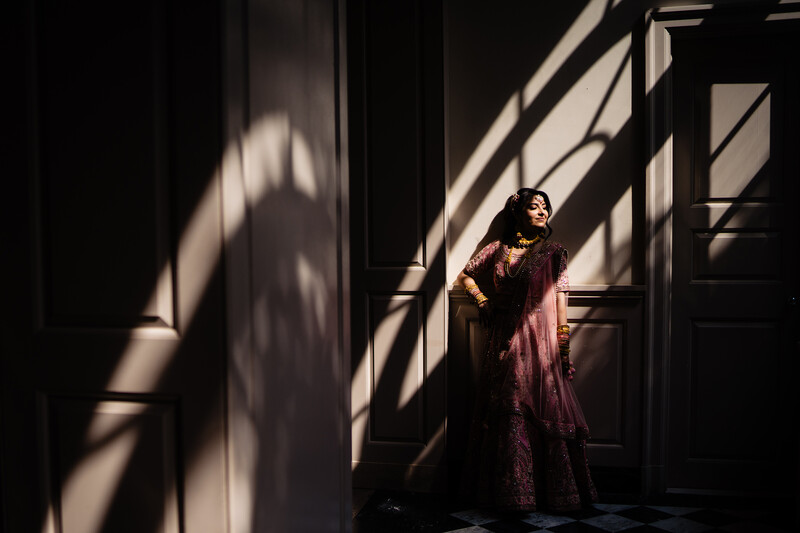 Woman in burgundy dress standing on stairs with dramatic window light creating geometric shadows across white walls.