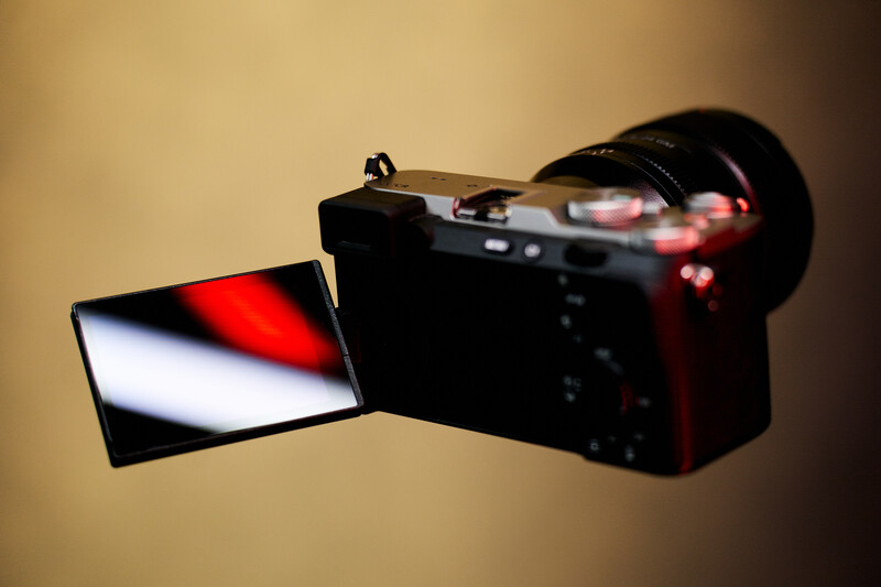 Mirrorless camera with articulating screen displaying red and white color gradient against warm beige background.