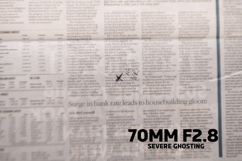 Newspaper page shot with 70mm f/2.8 lens showing severe ghosting artifacts across text and imagery.