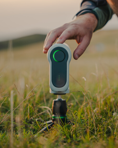 Hand holding a compact laser rangefinder device over grass in an open field.