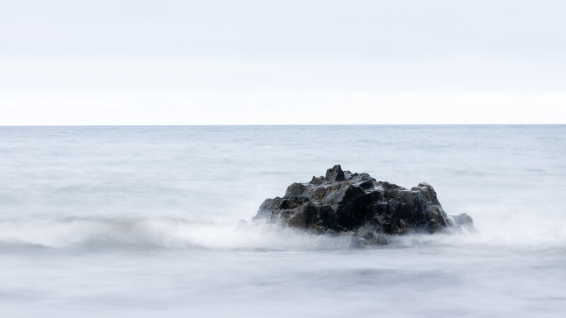 Dark rocky outcrop surrounded by churning white surf and mist in a long-exposure seascape.