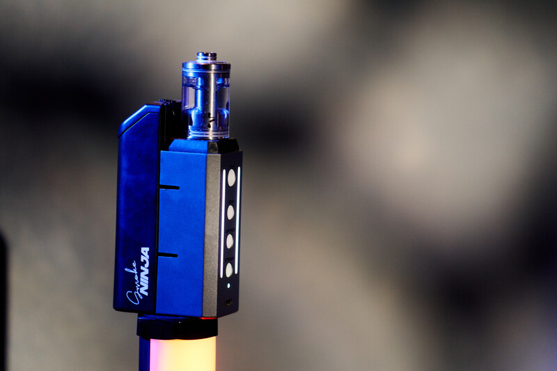 Blue and black vaping device with atomizer tank mounted on top, shot against blurred background.