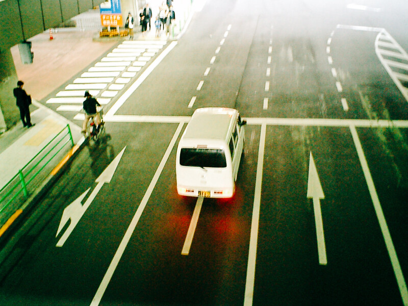 Aerial view of white van on multi-lane road with pedestrian crossing and people visible at street level.