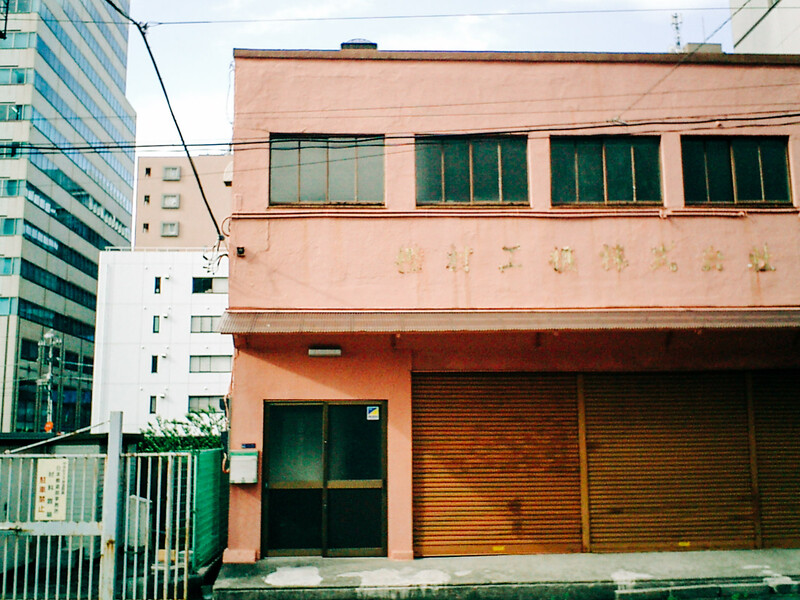 Weathered salmon-pink building facade with green-trimmed windows and closed orange roller shutters.