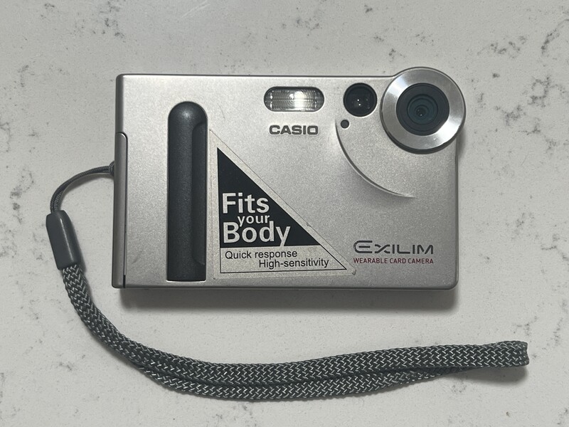 Casio Exilim compact digital camera with silver body and dark wrist strap on textured surface.