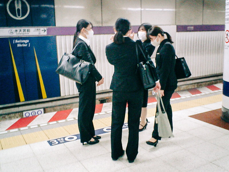 Four people in dark business attire standing in a subway station, wearing face masks.