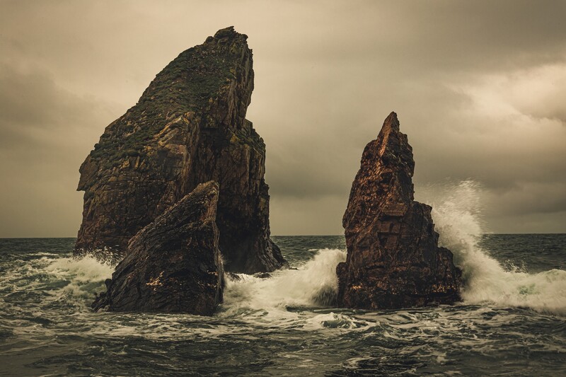 Two jagged rock formations rise from turbulent ocean waters under an overcast sky.