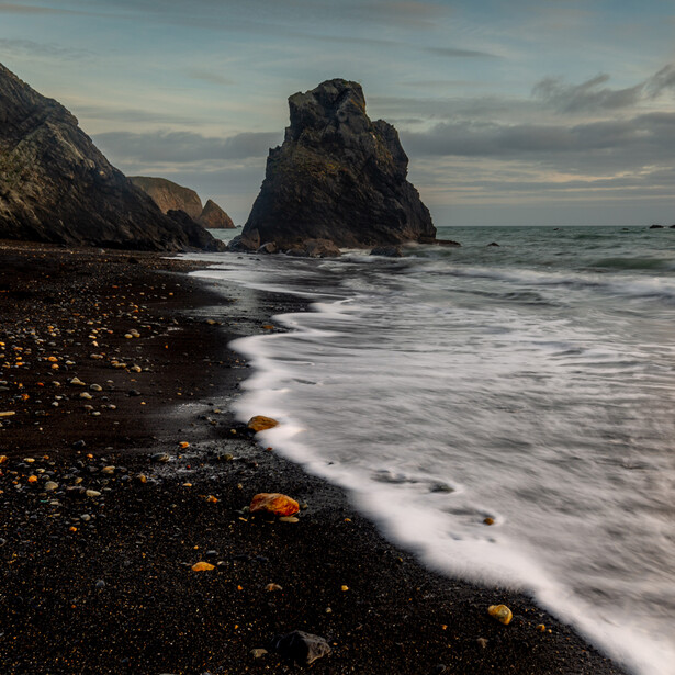 Rocky sea stack surrounded by waves on a dark pebble beach at dusk.