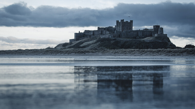 Medieval castle perched on a rocky headland, reflected in still tidal flats under moody gray sky.
