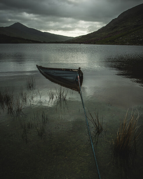 Wooden rowboat tethered in shallow water with mountains and overcast sky reflected in still lake.