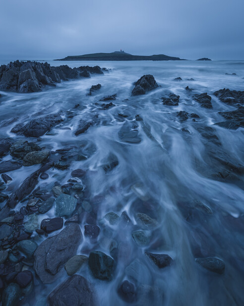 Rocky coastline at dusk with smooth water flowing between dark stones and distant islands on the horizon.