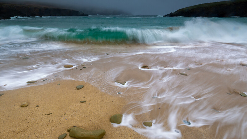 Turquoise wave with white foam breaking onto a sandy beach scattered with rocks.