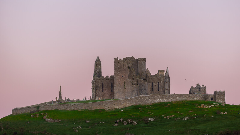 Medieval stone castle ruins on a green hillside against a pink twilight sky.
