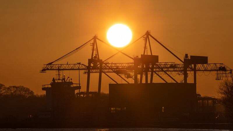 Silhouetted cargo ship and port cranes against a large sun at sunset or sunrise.