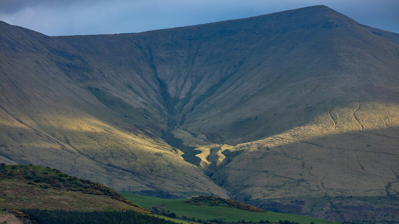 Mountain valley with dramatic shadows and a winding stream cutting through green pastures below steep slopes.