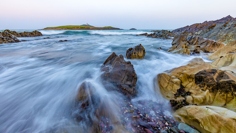 Waves crash over rocky outcrops at twilight, with a distant island visible across calm blue waters.