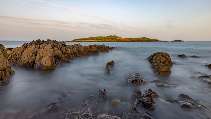 Rocky coastline with smooth water and distant lighthouse on an island at dusk.