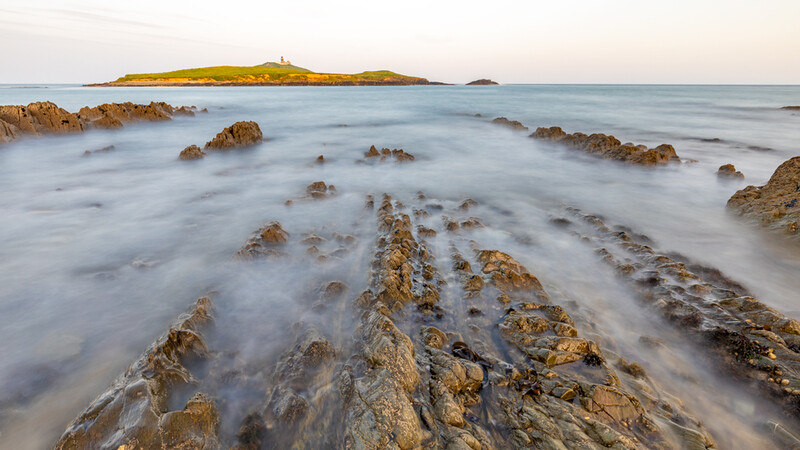 Rocky coastline with long-exposure ocean swells and a green island in the distance.