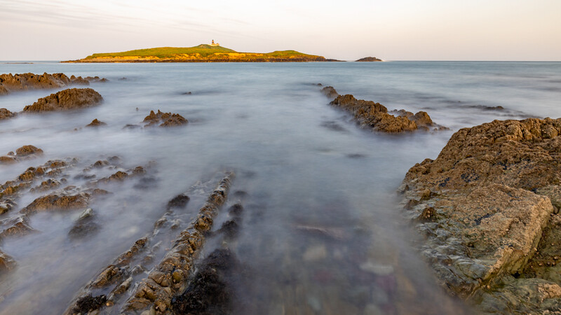 Rocky coastline with smooth water and distant island lit by golden hour light.