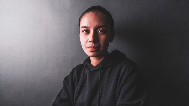 Young person in dark hoodie against a gray studio backdrop, lit from the side.