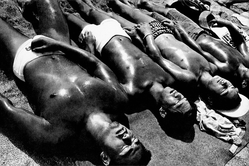 Multiple shirtless men lying on sand in bright sunlight, shot from above in black and white.