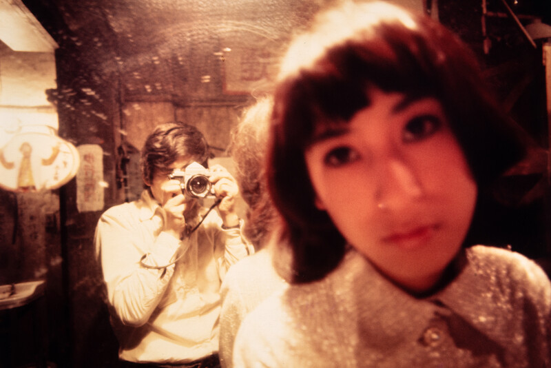 Photographer holding vintage film camera with subject in soft-focus foreground, warm color cast suggesting aged film stock.