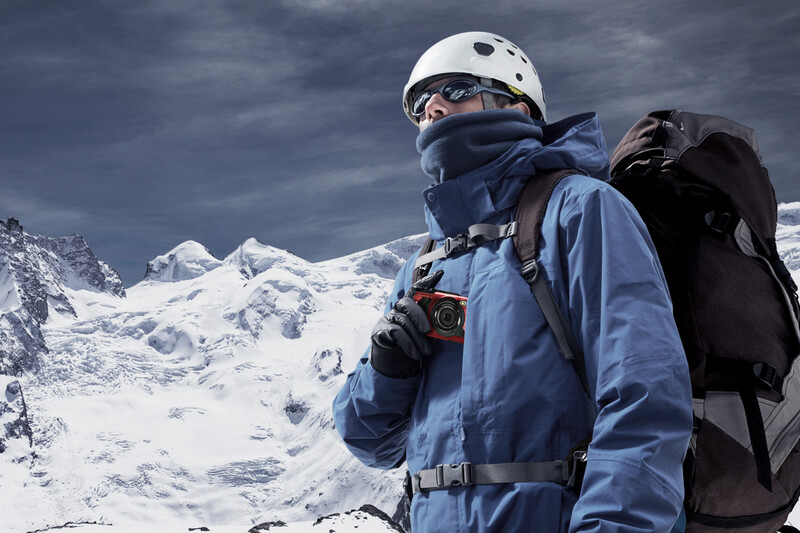 Mountain climber in blue jacket and white helmet stands against snow-covered peaks under dramatic sky.