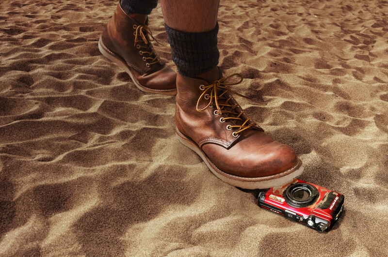 Person wearing brown lace-up boots standing on sand next to a toy car.