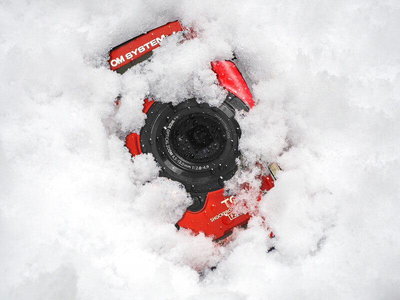 Red and black action camera partially buried in fresh snow, viewed from directly above.