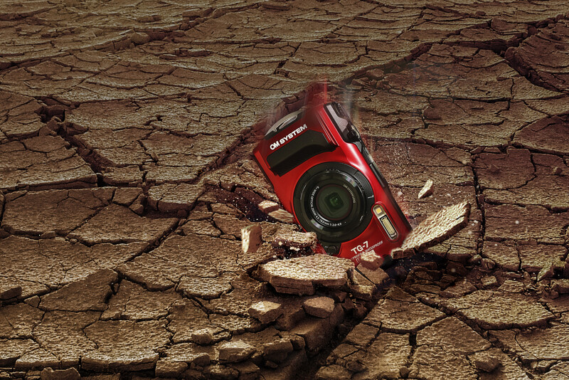 Red compact camera resting on cracked, dried earth with scattered twigs and debris.