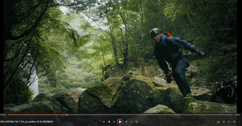 Hiker leaping across moss-covered boulders in a dense, verdant forest with a stream visible in the background.