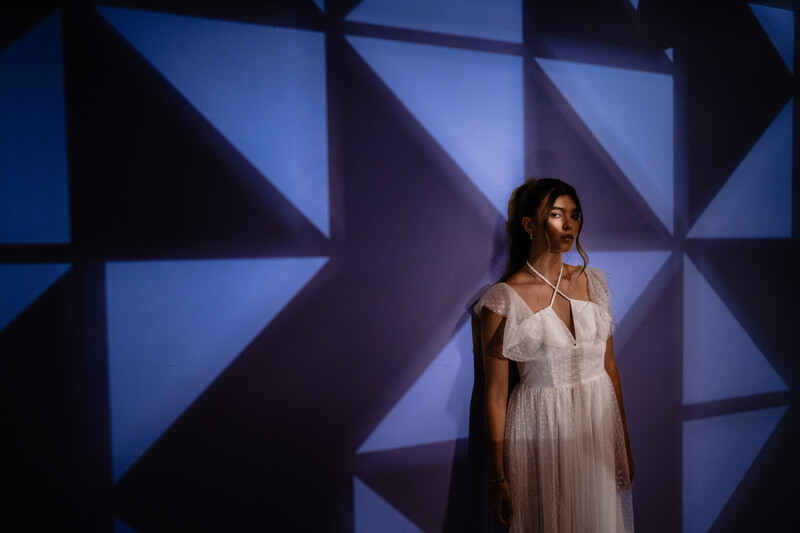 Woman in white dress standing against geometric blue and black backdrop with triangular light patterns.