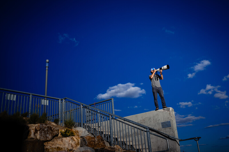 Man standing on concrete structure holding telescope toward sky against blue background.