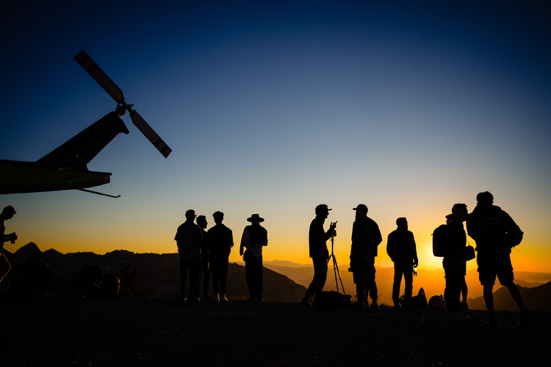 Silhouetted group of people standing beneath a helicopter against a golden sunset sky.