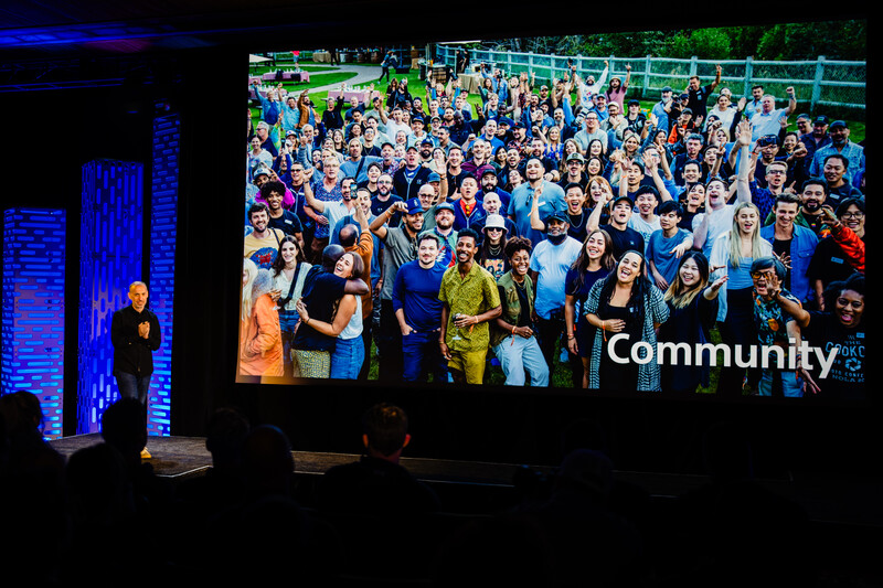 Presenter on stage left beside large screen displaying diverse group photo labeled 'Community'.