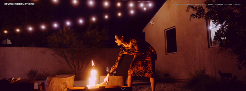 Person crouched by fire pit at night with string lights overhead and residential buildings in background.